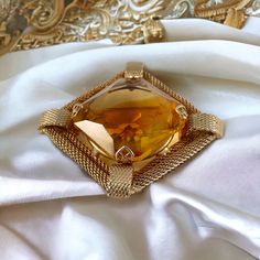 Elevate your style with this stunning vintage givre glass rhinestone and gold-plated mesh brooch, a true gem from the 1960s. The centerpiece boasts a large square-cut faceted glass stone, measuring just over 1" x 1". This mesmerizing stone showcases a captivating blend of light topaz with a deeper topaz hue at its center, all held securely in place by ornate prongs. Surrounding this remarkable stone is a square frame crafted from intricately detailed gold-plated mesh rope, featuring mesh half circles at each corner for added flair. While this piece is expertly constructed, it bears no signature, though it is reminiscent of similar creations by Hobe, known for their gold mesh designs. Resting in very good vintage condition, this brooch promises to stand the test of time. With its unique and Luxury Yellow Gold Brooches For Parties, Gold Brooches With 17 Jewels For Evening, Gold Jeweled Brooches For Formal Occasions, Formal Gold Jeweled Brooches, Elegant Yellow Brooches For Party, Elegant Yellow Party Brooch, Elegant Yellow Party Brooches, Vintage Yellow Gold Brooches For Party, Elegant Gold Jeweled Brooches