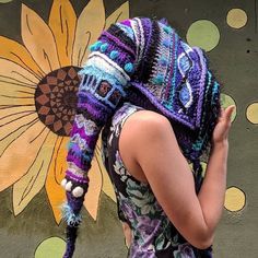 a woman wearing a crocheted hat and scarf standing in front of a flower painted wall