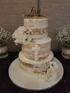 a three tiered cake with white flowers on the bottom and gold lettering that says first commution