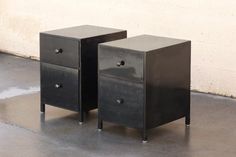 two black nightstands sitting next to each other on top of a cement floor in front of a white wall