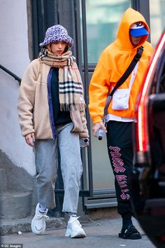 two people are walking down the street in front of a car and one is wearing an orange jacket