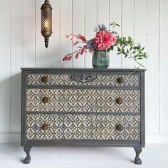 an old dresser with flowers on top and a lamp hanging from the wall behind it