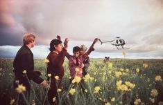 three people in a field with a helicopter flying over the top of them and one person holding up his hand