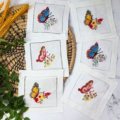 four embroidered napkins with colorful butterflies on them next to green leaves and straw basket