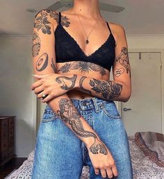 a woman with tattoos on her arm and chest standing in front of a bed wearing jeans