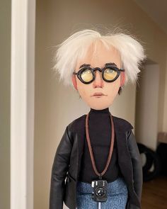 a doll with white hair wearing glasses and a black jacket is standing in front of a door