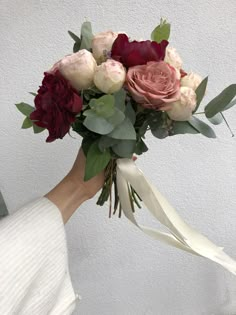 a woman holding a bouquet of flowers in her hand with white ribbon around it and greenery on the side