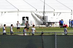 a group of children playing soccer on a field with people watching from behind a fence