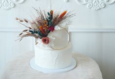 a white cake with flowers and feathers on top sits on a table in front of a wall