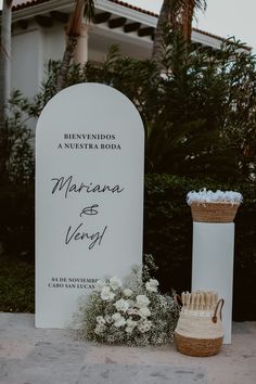 a white sign sitting next to a vase with flowers in it and a basket on the ground