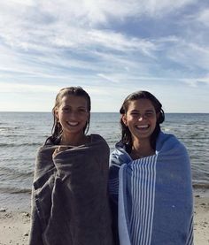 two girls wrapped in towels on the beach by the ocean, one is smiling at the camera