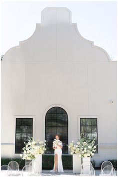 a woman standing in front of a white building with chairs and flowers on the ground