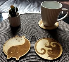 two cat coasters sitting on top of a table next to a cup and plant