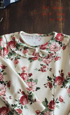 a white shirt with red flowers on it sitting on top of a wooden floor next to a pair of glasses