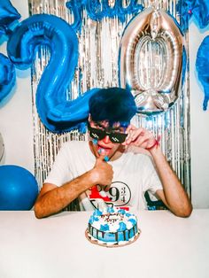 a man sitting at a table with a cake in front of him and balloons behind him