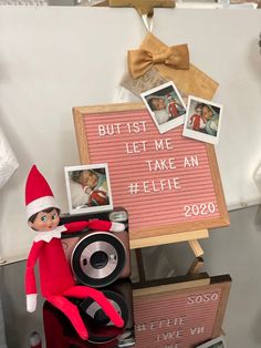 an elf sitting next to a sign with photos on it and a camera in the foreground