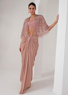 Elevate your style with the stunning Pastel Pink Draped Skirt with Embellished Cape, that effortlessly blends tradition with contemporary elegance. The pink cape features a sophisticated kimono sleeve , adorned with intricate florence embroidery using sequins, crystal tassels, cutdana, and bead highlights, adding a touch of opulence. Paired perfectly with a sequin jaal embellished blouse, it offers a dazzling appeal, making you the center of attention. Completed with a draped skirt, designed for a flattering silhouette, exudes grace and charm, ensuring comfort and sophistication in every movement. Ideal for, this ensemble is a timeless addition to your wardrobe, exuding both modernity and tradition in every detail. Composition : Cape - Organza ,Skirt - Platinoir Crepe Care: Dry Clean Only Drape Sarees, Embroidered Cape, Pink Cape, Pink Kimono, Indian Wedding Wear, Draped Skirt, Embellished Blouse, Kimono Sleeves, Indian Designer Outfits