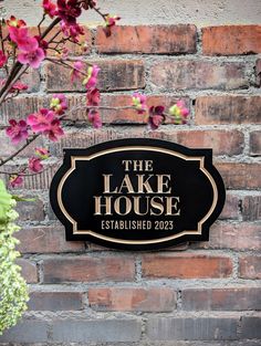 the lake house sign is next to some pink flowers on a brick wall and there are purple flowers in front of it