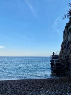 a man standing on the edge of a cliff next to the ocean with his boat in the distance