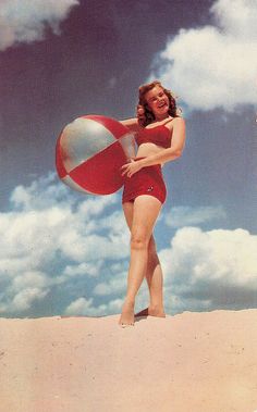 a woman in a red bathing suit holding a beach ball on the sand under a cloudy blue sky