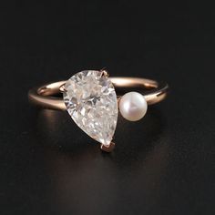 an engagement ring with a pear shaped diamond and a single pearl on the side, sitting on a black surface