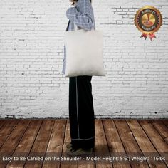 a woman holding a white tote bag on top of a wooden floor next to a brick wall