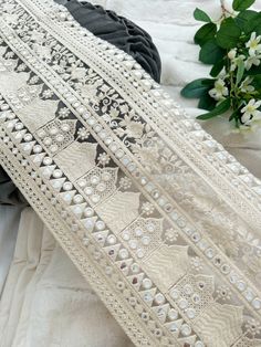 white lace with pearls and beads on it next to a bouquet of flowers in the background