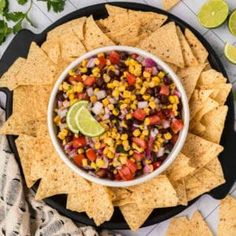 a black plate topped with corn salsa and tortilla chips next to lime wedges