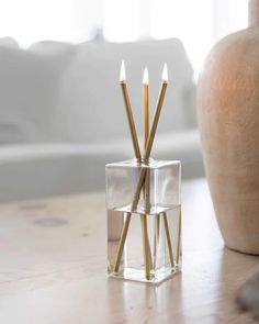 three candles sitting in a glass block on a table next to a vase with a white couch behind it