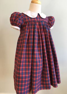 "This homemade dress was very nicely constructed and finished. Made from a poly/cotton blend, it is a woven tartan in red, blue, black, green and white. The waistline is high - an Empire style. The front bodice is finished with a very narrow bias self piping. The skirt below is gathered and mid-calf length. Sleeves are short and very puffed. Cuff is white and finished with self piping. Collar is likewise white with piped edges. It is round and wide. Back bodice sits lower than front and closure Fitted Plaid Dress For Fall In Preppy Style, Fitted Plaid Dress For Fall, Preppy Style, Fitted Plaid Preppy Dress For Fall, Plaid Smocked Short Sleeve Dress, Classic Fitted Plaid Dress For Fall, Short Sleeve Plaid Smock Dress, Fitted Plaid School Dress, Fitted Plaid Dress For School, Fitted Red Plaid Dress For Fall