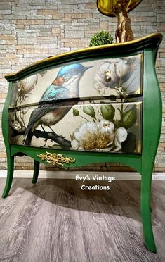 a green painted dresser with flowers and birds on it's drawers, sitting in front of a brick wall