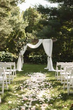 Garden wedding ceremony with a DIY wooden arch draped in white fabric and adorned with flowers, set in a lush, green outdoor space with white chairs and a flower petal aisle. Outdoor Wedding Ceremony And Reception, Garden Wedding Seating Ideas, Wedding Alter Outside, Elegant Backyard Wedding Ceremony, Outside Backyard Wedding Ideas, Spring Garden Wedding Ceremony, Wedding Ceremony Isles, Outdoor Garden Ceremony