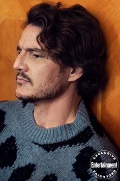 a man with long hair wearing a sweater and looking off to the side in front of a wood paneled wall