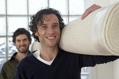 two men are smiling and one is carrying a mattress