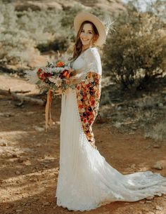 a woman in a long white dress and hat holding a flower bouquet on her wedding day