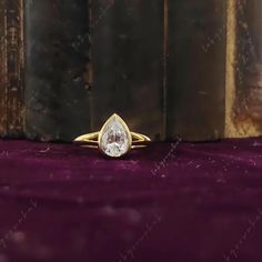 a yellow gold ring with a pear shaped diamond on the middle, sitting on a purple velvet surface