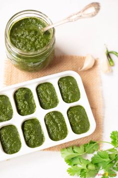 green pesto sauce in an ice tray next to garlic and parsley on a white surface