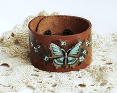 "A soft camel brown leather cuff decorated with a large butterfly stud, accented in turquoise blue and surrounded by tiny painted blue flowers sprinkled among golden-green leaves! A few select flowers sparkle with the help of little blue Swarovski crystal rhinestones for a touch of glam! The cuff is sure to fit most wrists as it is adjustable with two sturdy snap size options! An artful addition to your wardrobe! Adjustable Soft Leather Cuff, 9\" long, 1 1/2\" wide with two snap size options, wi Adjustable Brown Jewelry For Spring, Brown Jewelry For Spring Gift, Spring Brown Jewelry Gift, Spring Gift Brown Jewelry, Hand Painted Butterfly, Painted Butterfly, Hand Painted Candles, Golden Green, Leather Cuff Bracelet