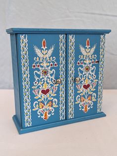 a blue cabinet with ornate designs painted on it's doors and handles, sitting on a white surface