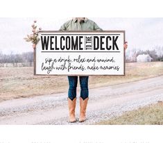 a man holding a sign that reads welcome to the deck, and standing on a dirt road