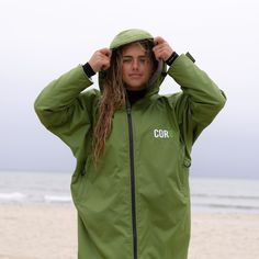 a man with long hair wearing a green jacket on the beach and covering his ears