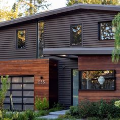 a modern house with wood sidings and black garage doors on the front, surrounded by greenery