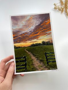 a hand holding up a card with an image of a sunset in the sky over a field