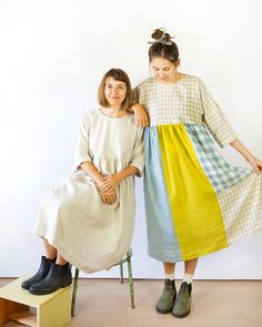 two women in dresses sitting on stools with their arms around each other's shoulders