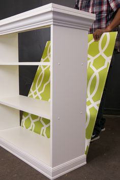 a white book shelf with green and white wallpaper on the top, in front of a man standing next to it