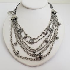 Fun rocker bib necklace with 6 strands of silver tone metal chains attached with inscribed bell shaped end covers. The chains are interspersed with glass beads of various shaped and sizes with a metallic finish. The embellishment just past the end covers dangles on one side and has a larger silvered jewel on the other. Very eye catching with the kinetic movement and shine.  The bib is approximately 8" by 3". The necklace length is approximately 20". Previously owned. Appears to be in good condit Silver Multi-strand Metal Chain Necklace, Multi-strand Silver Chain Necklace, Silver Beaded Chain Layered Necklace, Silver Layered Beaded Chain Necklace, Silver Bib Necklace With Chain In Costume Jewelry Style, Silver Bib Necklace With Chain For Costume Jewelry, Silver Chain Bib Necklace For Costume Jewelry, Silver Multi-strand Metal Layered Necklace, Silver Multi-strand Beaded Chain Necklace