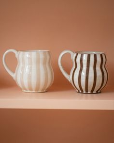 two white and brown striped coffee mugs on a pink shelf next to each other