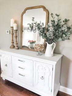 a white dresser topped with a vase filled with flowers next to a mirror and candle