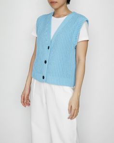 a woman wearing a blue knitted vest and white pants standing in front of a wall