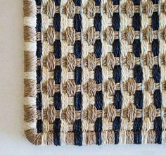 a woven wall hanging with black and tan strips on it's side, next to a white wall